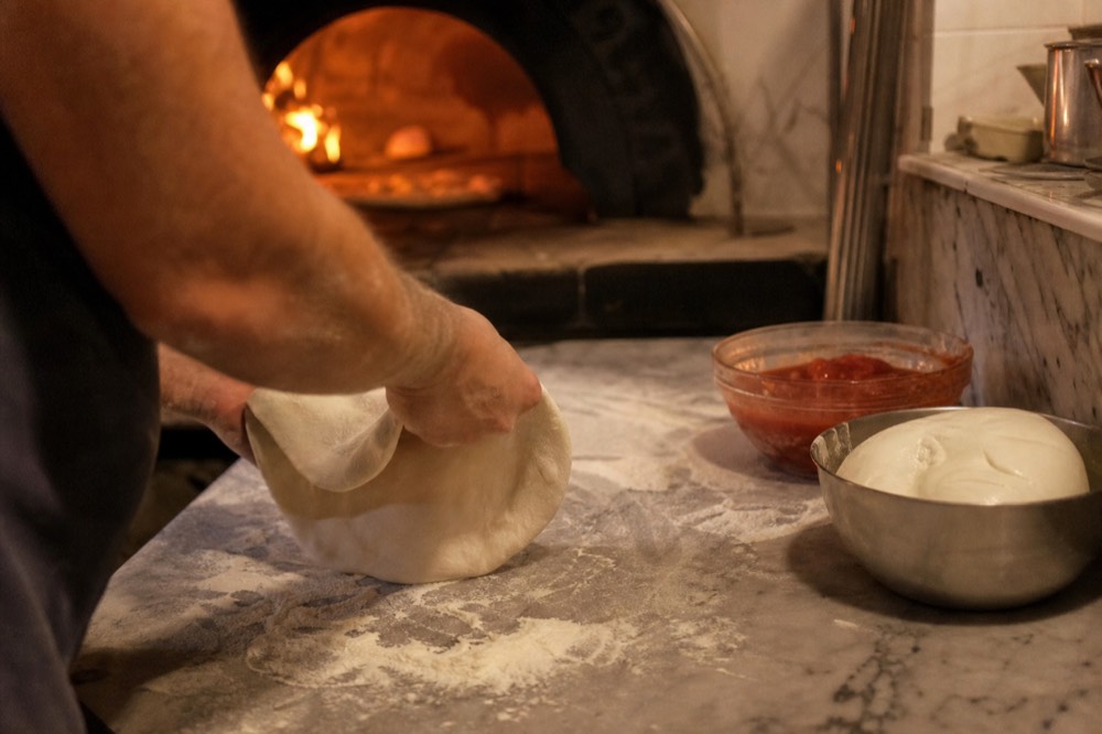 Hands stretching fresh pizza dough on a floured bench beside a wood-fired oven