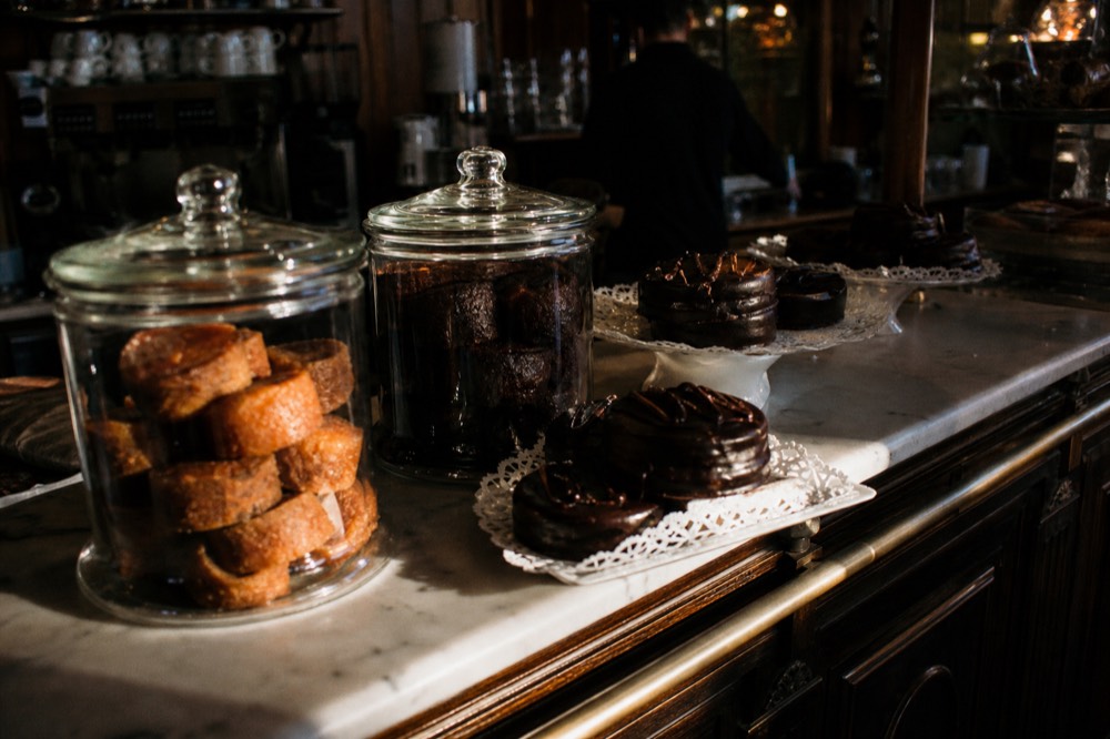 Marble pastry counter stacked with rum-soaked cakes in glass jars and dark chocolate rounds on paper doilies under warm shop light