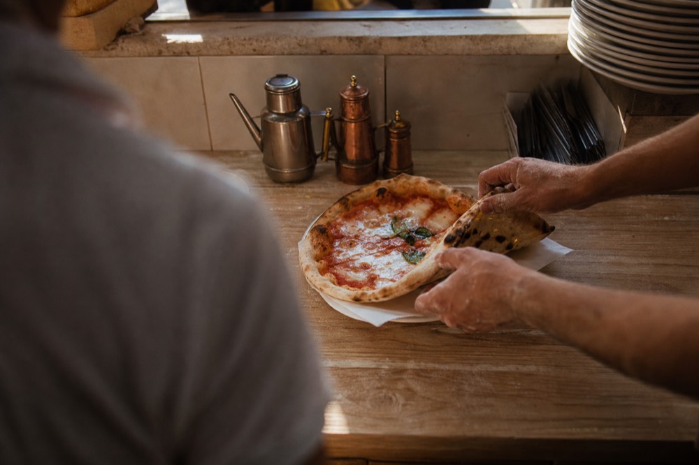 A pizza being folded in half then quarters on a wooden counter and wrapped in plain white paper by flour-dusted hands