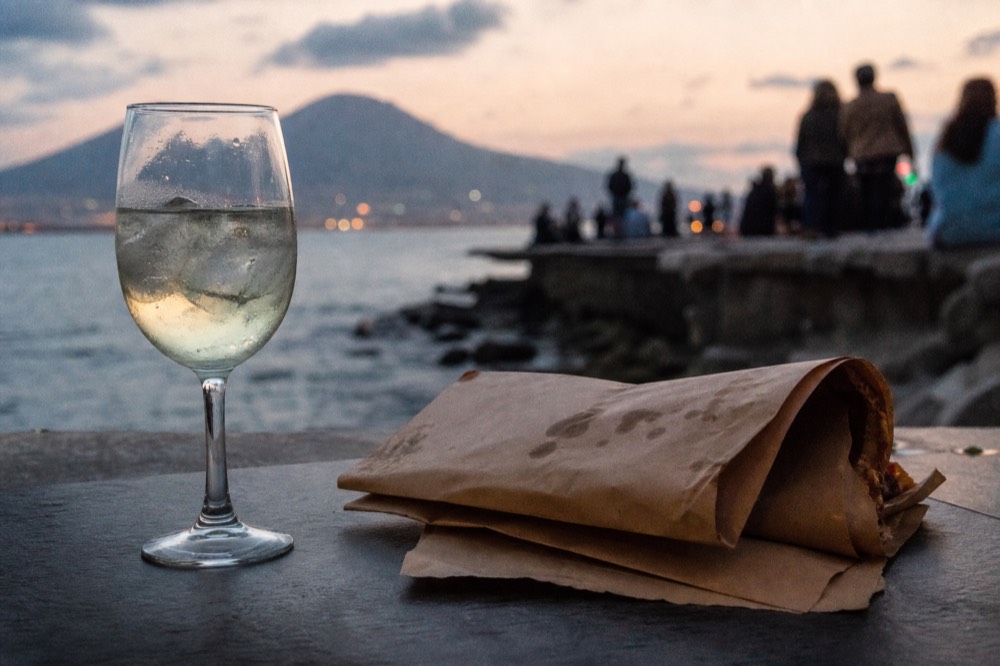 A cold spritz in a stemmed glass beside a folded paper-wrapped pizza on an outdoor table at dusk in Naples