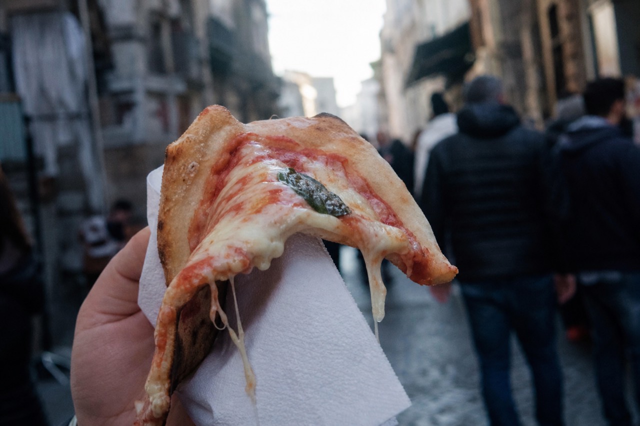 A folded quarter of pizza held in a paper napkin at street level, cheese stretching, blurred alley crowd behind.