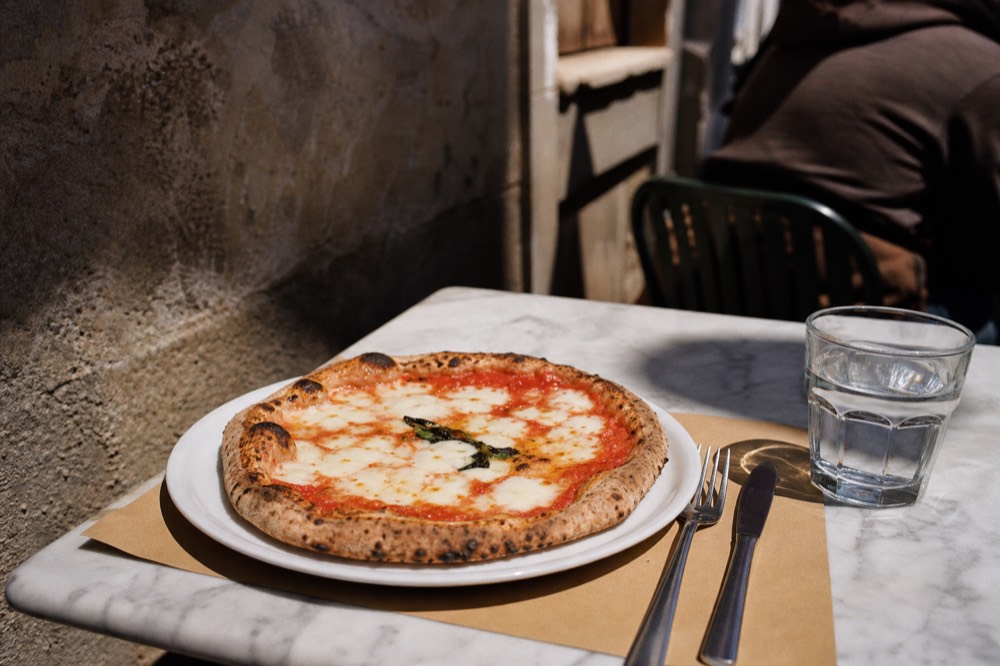 A plain margherita pizza on a simple white plate with a paper placemat, cutlery and a glass of water on a marble table