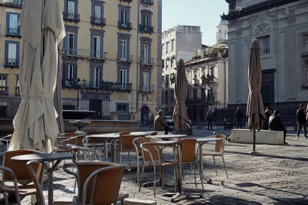 Busy Naples piazza terrace at midday with empty café tables, folded parasols and worn paving stones, no signage visible