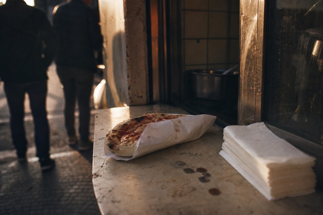 Coins and a folded paper-wrapped pizza on a worn counter beside a stack of white napkins at a street window.