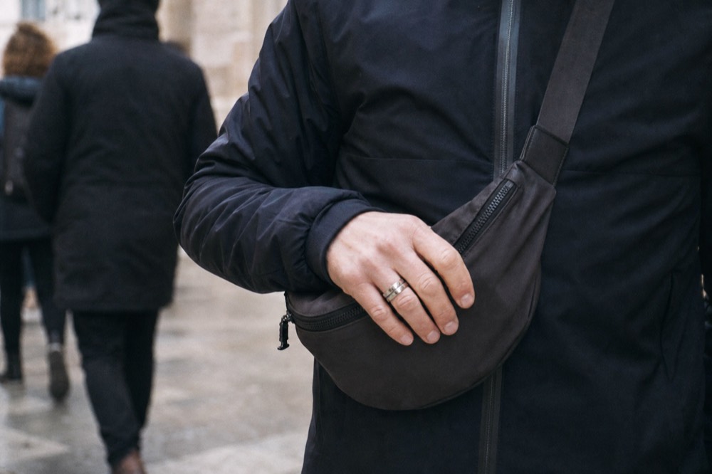 A crossbody bag worn tight across a jacket in a crowded street, one hand resting on the zipped flap, blurred passers-by behind