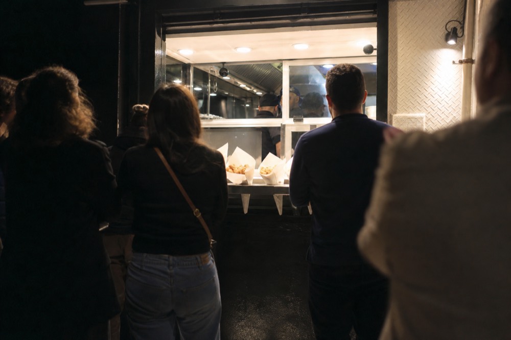 Backs of a small group gathered at a lit street food counter at night, paper cones of fried food resting on the ledge