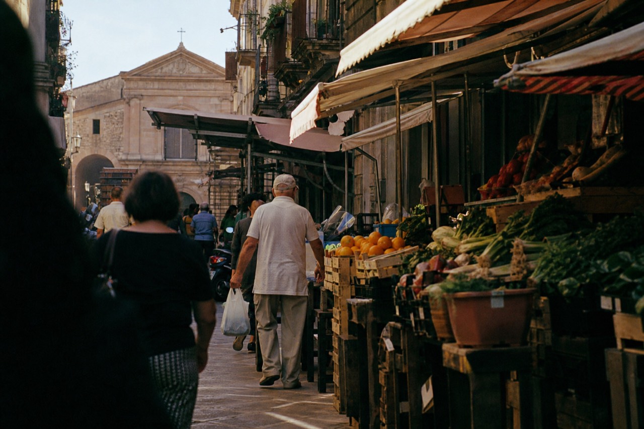 Busy market street in daylight with shoppers carrying bags, produce stalls under awnings, parked scooters and an old church wall at the far end