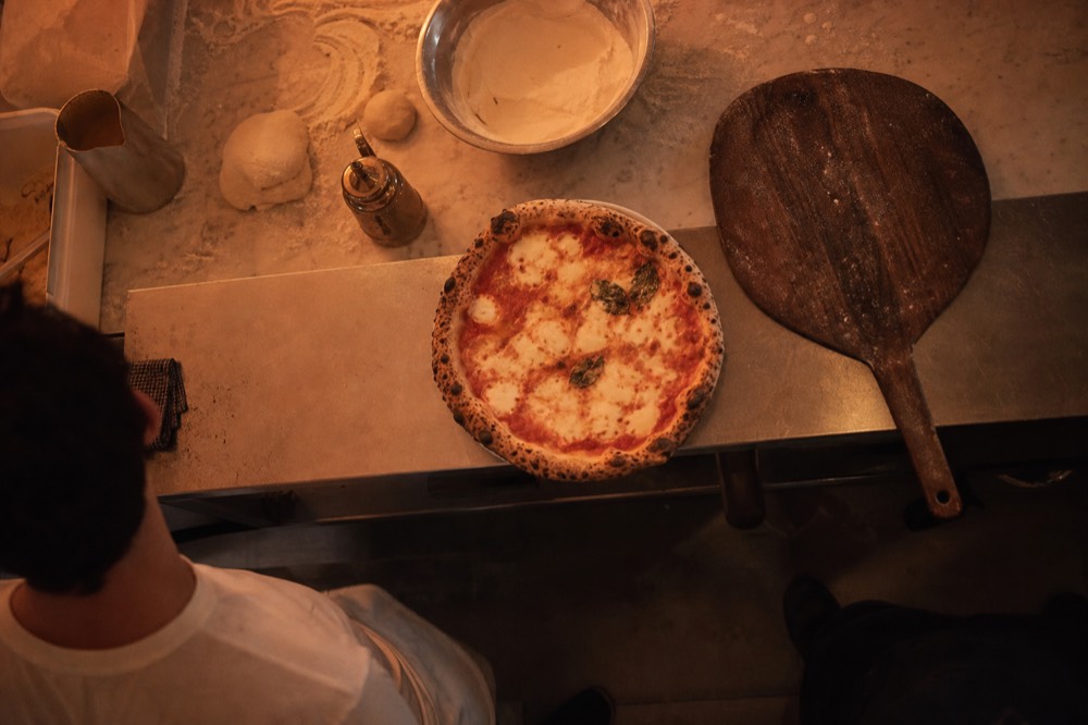 Overhead view of a bubbling charred margherita pizza on a steel counter beside a wooden peel and a flour-dusted marble bench