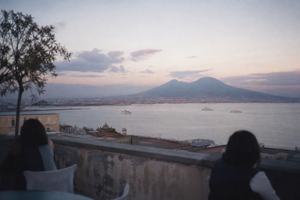 Wide view across the bay at dusk from a rooftop terrace, ferries crossing calm water, a volcano silhouette low on the horizon