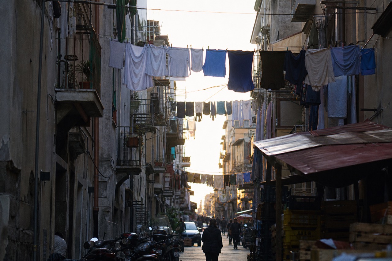 Laundry strung between tall balconied buildings above a crowded alley at golden hour, scooters and stacked market crates on the pavement below