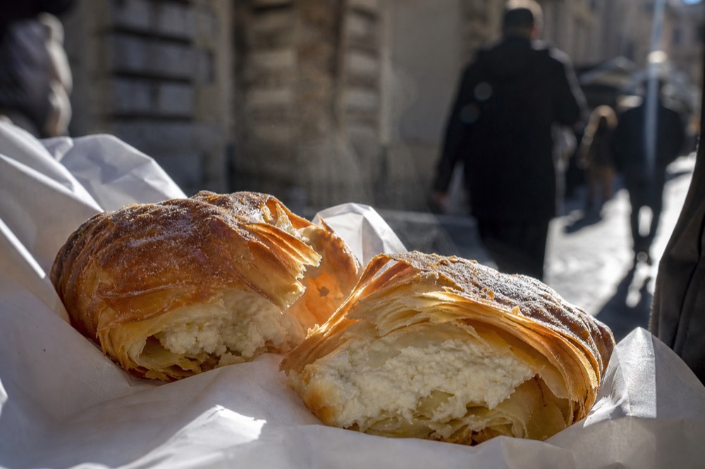 Close-up of a freshly baked layered pastry broken open with steam rising, flaky ridges and ricotta filling visible on white paper