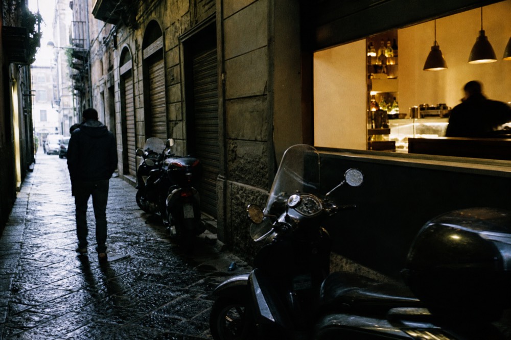 Narrow Naples street at early morning with shuttered shops, a lit corner bar counter glowing inside, scooters parked along wet cobblestones