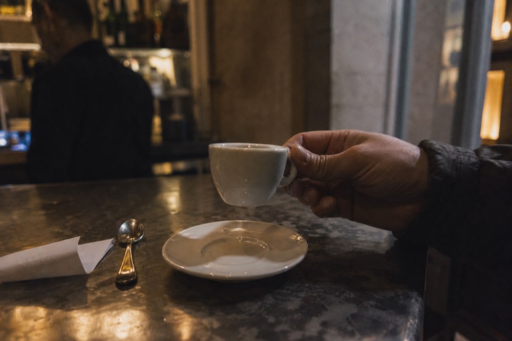 Hands lifting a small white espresso cup from its saucer on a zinc bar counter, sugar spoon and folded receipt resting beside it
