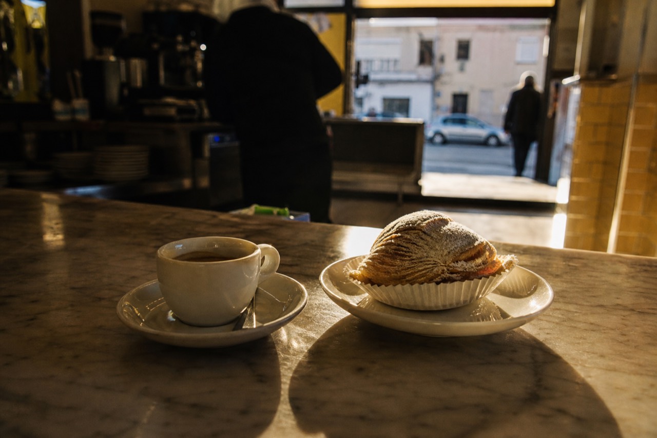 Espresso cup and a shell-shaped ricotta pastry on a marble bar counter at dawn, warm light coming in from the street outside