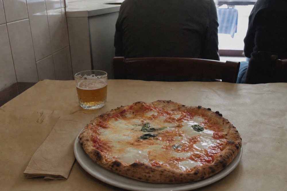 Simple margherita pizza beside a small glass of beer on a paper-covered table in a plain neighbourhood pizzeria