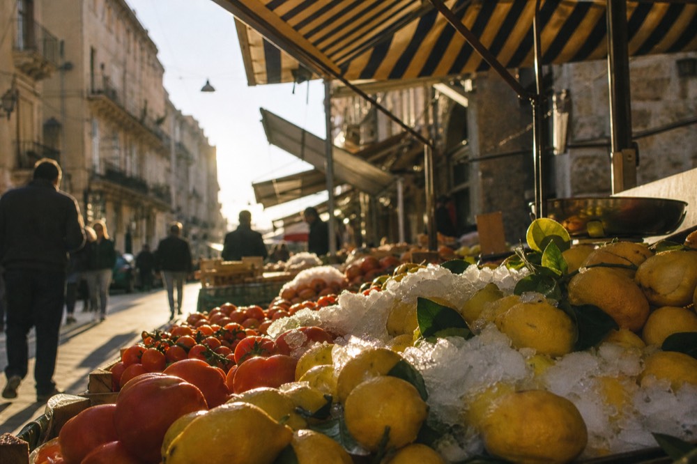 Market stall stacked with lemons, tomatoes and crushed ice under a striped awning on a busy narrow shopping street
