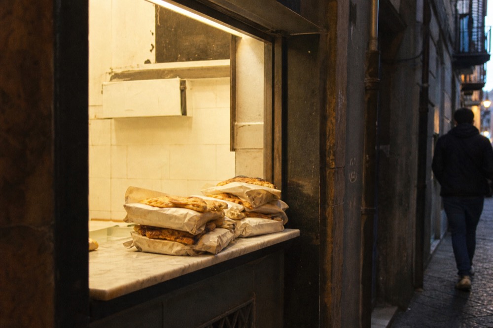 Street window of an old pizzeria on a narrow Naples lane with paper-wrapped folded pizzas stacked on a marble counter