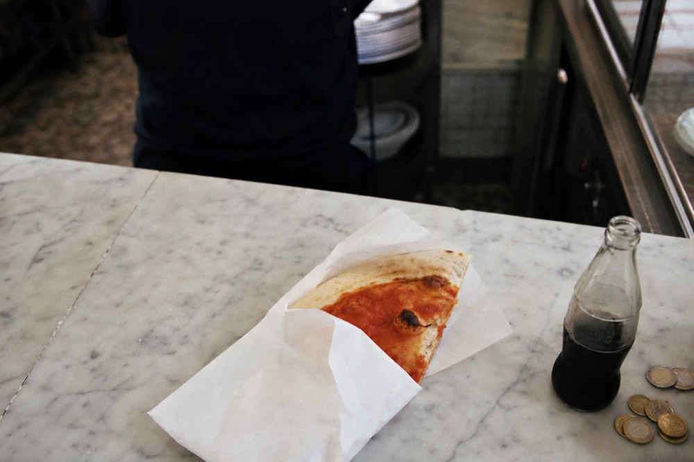 Overhead view of a marble counter with a folded paper-wrapped pizza, a glass bottle of cola and coins beside it