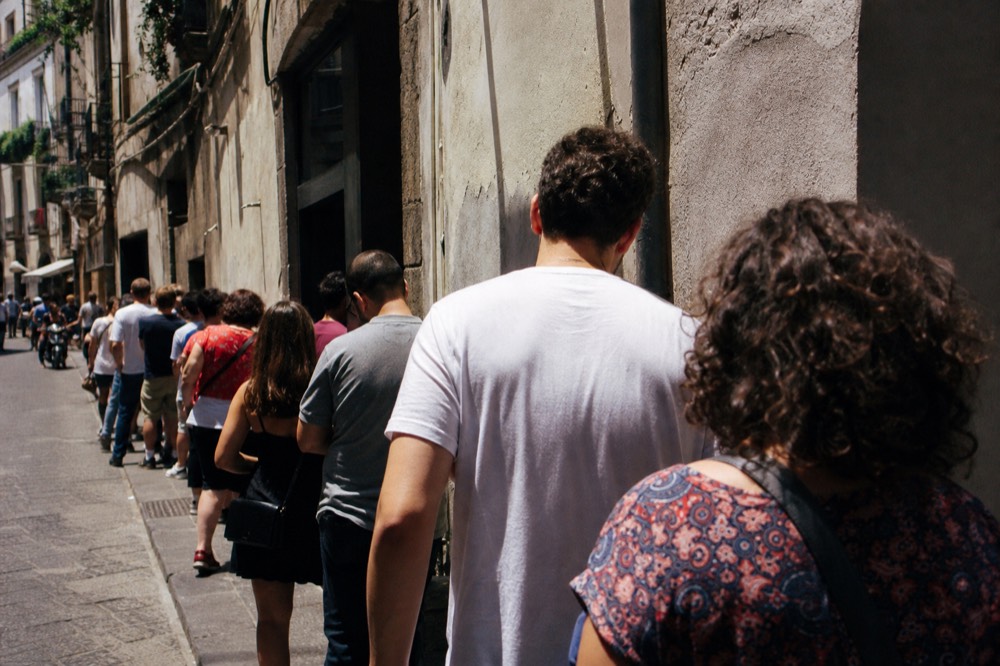 Long line of people waiting along a sunlit Naples pavement outside an old pizzeria doorway, seen from behind at street level