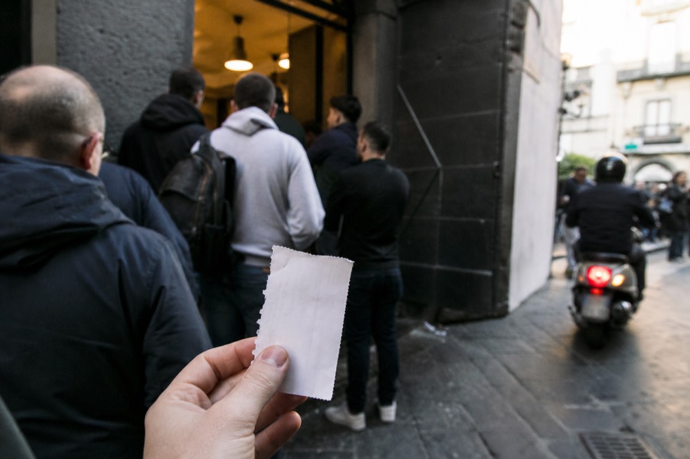 Numbered paper ticket held in a hand outside a busy pizzeria door, blurred crowd and scooter traffic in the background