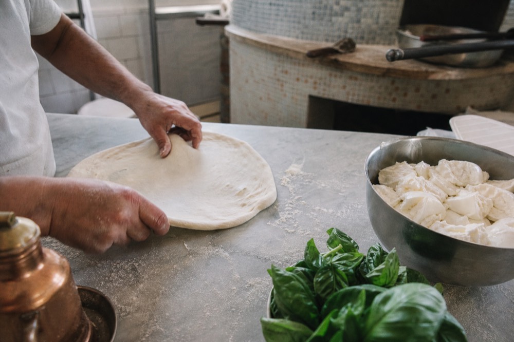 Hands stretching a pale disc of pizza dough on a floured marble bench beside a bowl of torn mozzarella and fresh basil