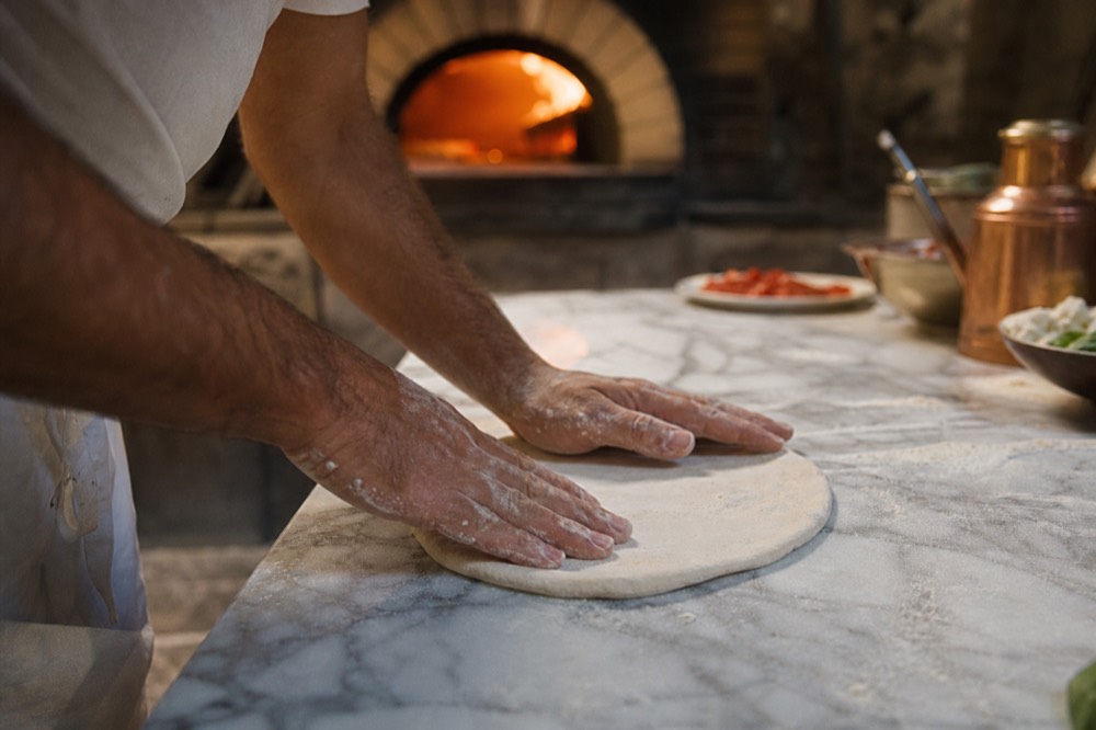 Pizzaiolo's flour-dusted hands pressing a dough ball outward on a marble bench, wood oven glowing out of focus behind