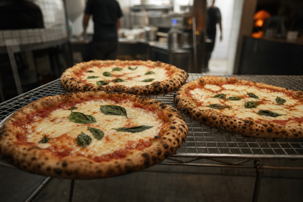Three whole margheritas cooling on a steel rack under warm pizzeria light, rims blistered dark, basil leaves scattered across each