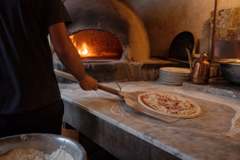 Hands sliding a raw pizza onto a wooden peel beside a flour-covered marble counter in a small traditional bakery workshop