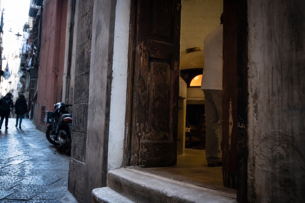 A worn wooden pizzeria doorway on a narrow Naples lane, stone step rubbed smooth, warm light spilling from inside