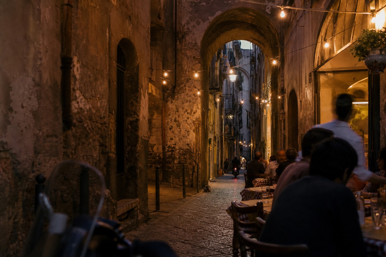 A narrow Naples alley of old stone arches at dusk with pizzeria tables spilling onto the cobbles and warm light overhead.