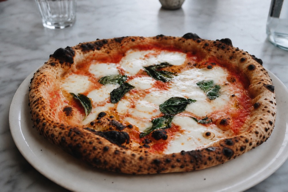 Blistered margherita on a plain white plate, charred cornicione bubbles, basil wilting into molten mozzarella, marble table, overhead daylight, no branding