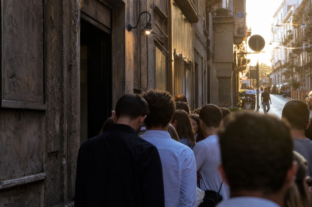 Crowd of waiting diners spilling onto a narrow cobbled Naples street outside a pizzeria doorway at dusk, shot from behind, faces away
