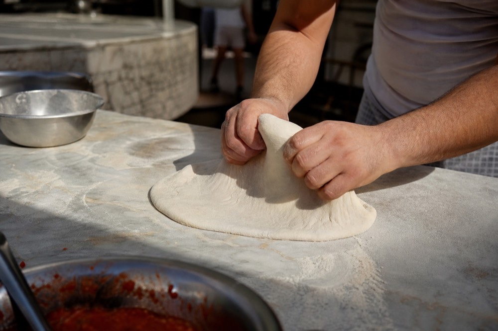 Pizzaiolo's hands stretching a pale dough ball on a floured marble counter, flour dust in low light, forearms only, no faces