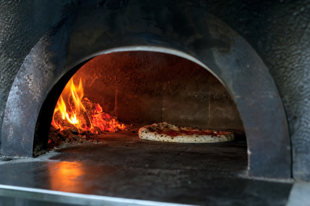 Interior of a wood-fired dome oven, orange embers banked to one side, a pizza puffing in seconds against blackened firebrick