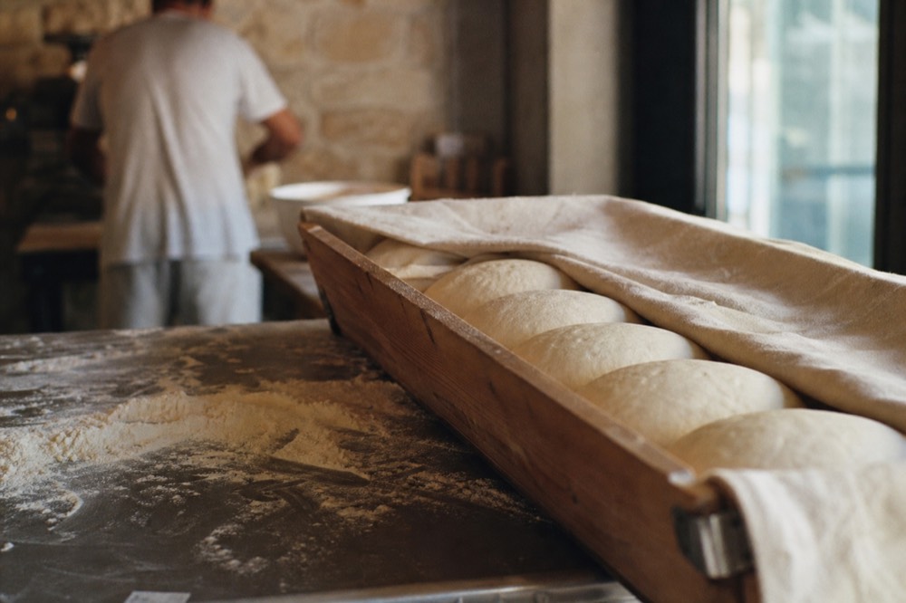 Row of dough balls resting in a wooden proofing box under cloth, soft window light, flour scattered on a steel bench