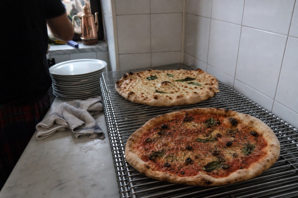 Two pizzas cooling on a steel rack beside a stack of white plates and a folded cloth in a tiled kitchen corner