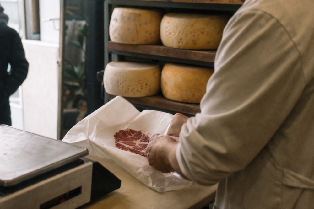 Hands wrapping a paper parcel of sliced cured meat on a scale-topped counter, wheels of cheese stacked on shelving behind