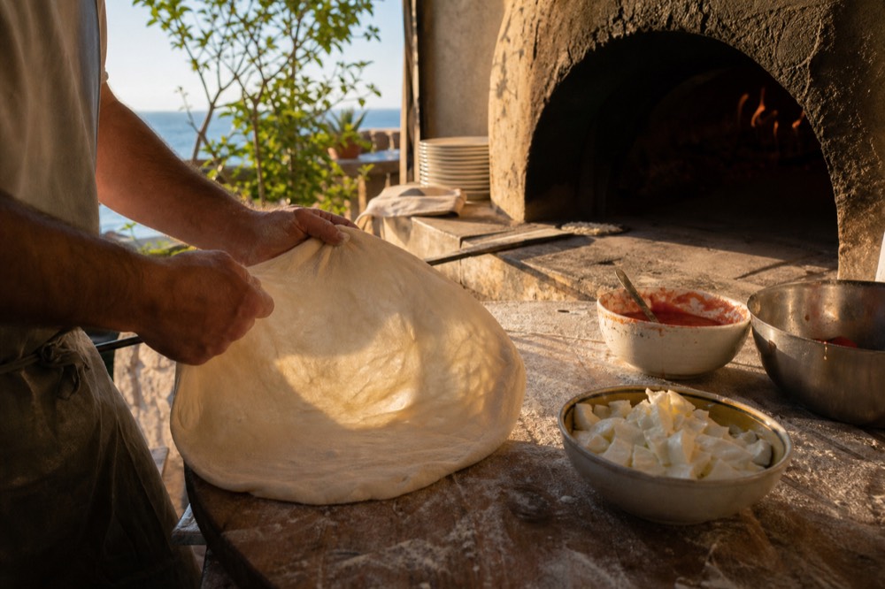 Floured wooden work surface with hands stretching a pale dough disc, bowls of tomato and torn cheese waiting beside a wood oven mouth