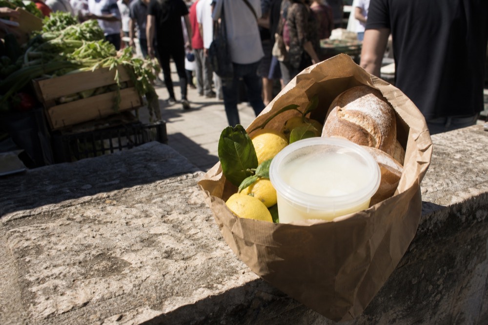 Paper shopping bag on a stone ledge holding lemons, bread and a whey-filled tub, market crowd blurred in the background