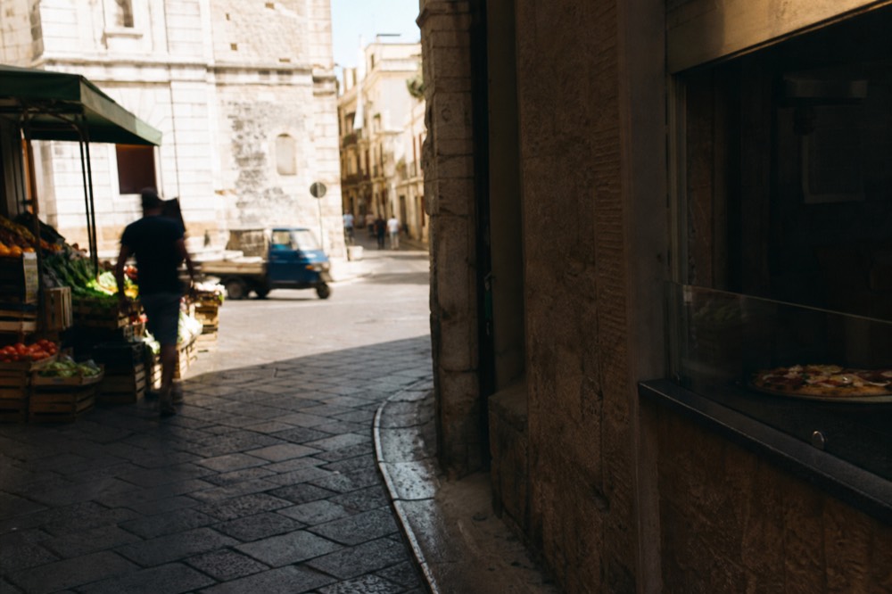 Street corner where a narrow historic lane meets a wider boulevard, pizza window on one side and market crates on the other, midday shadow