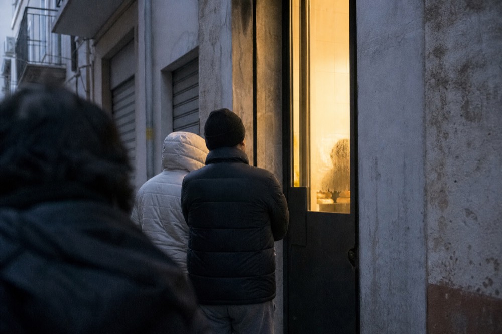 Early morning queue outside a small bakery doorway on a narrow side street, warm interior light and steam on the glass, closed shutters opposite