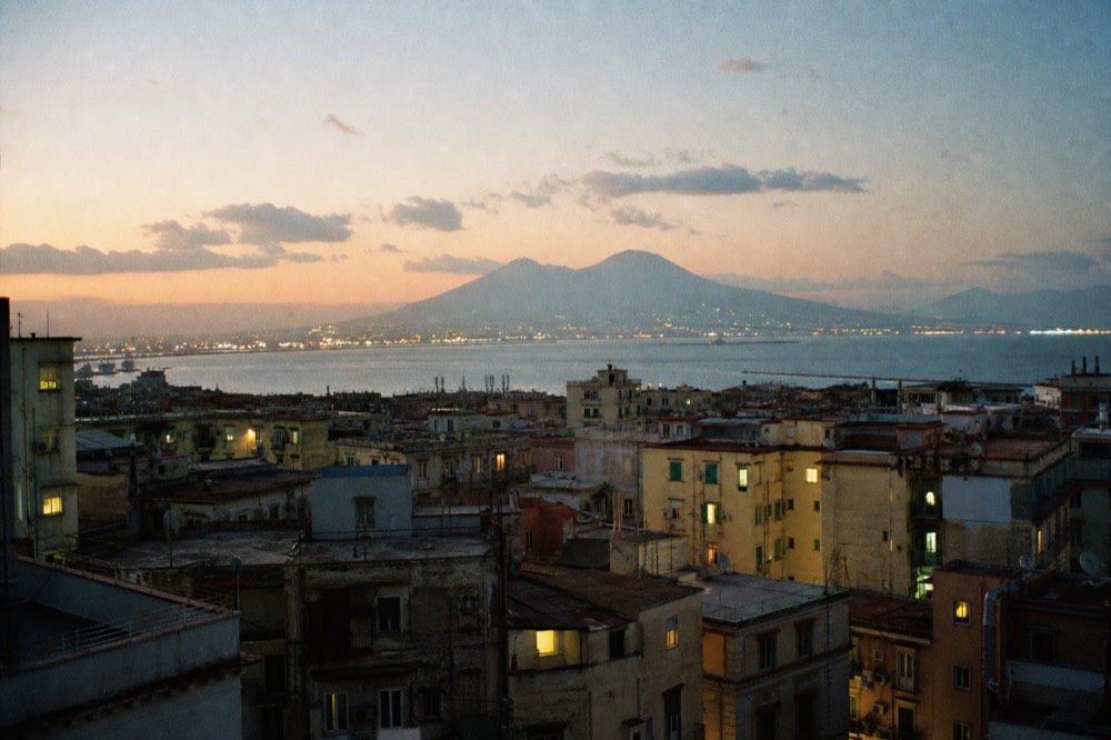 Rooftop-level view across dense Naples housing toward the bay at dusk, lit windows and a hazy volcano outline on the horizon