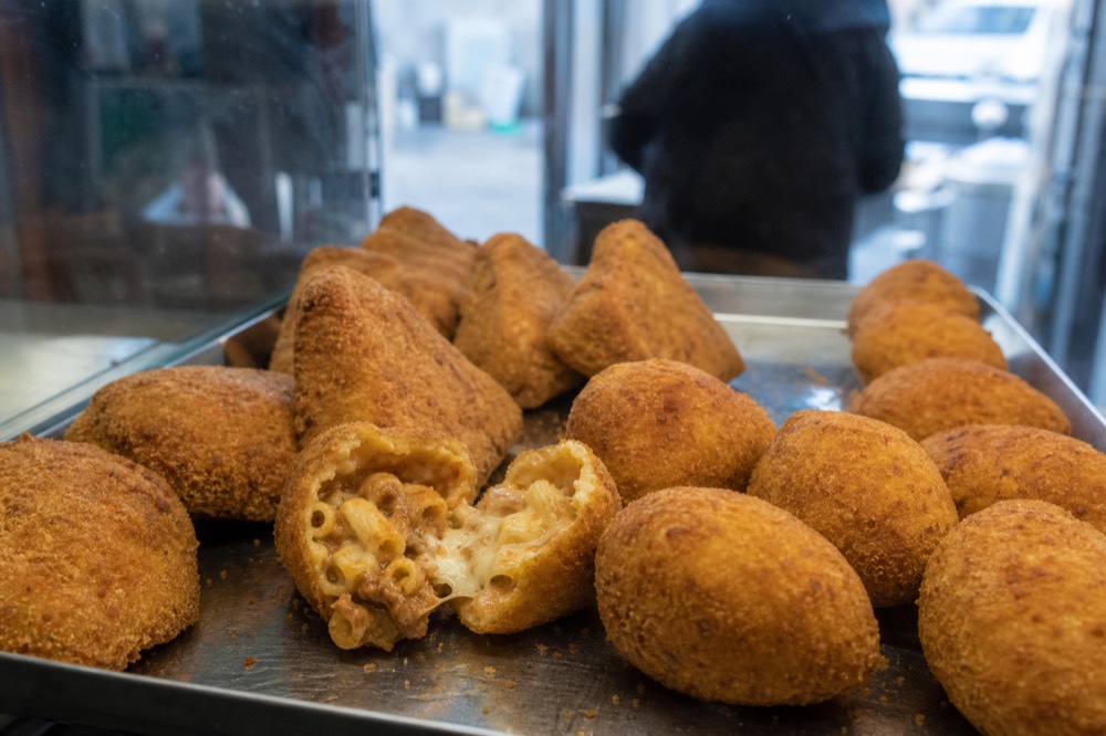 Steel tray of fried pasta cakes and potato croquettes under counter glass, one broken open showing creamy pasta and melted cheese inside