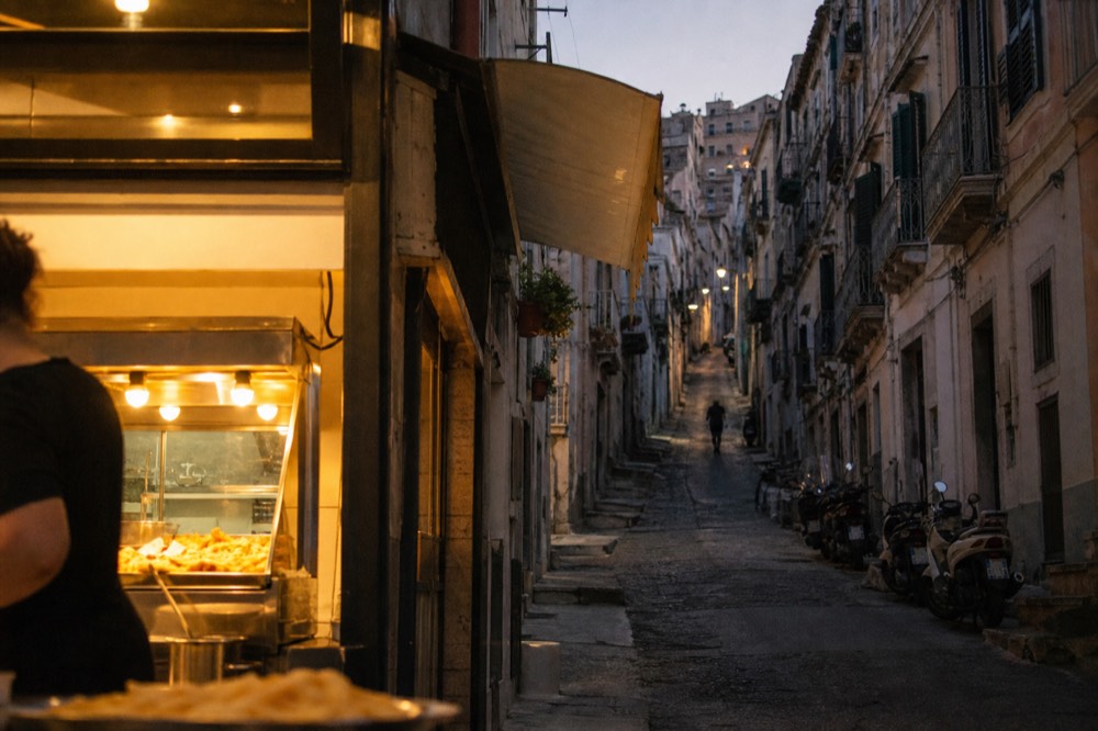 Steep residential lane climbing between tall shuttered buildings, a small fry counter glowing at the bottom, scooters parked along one wall