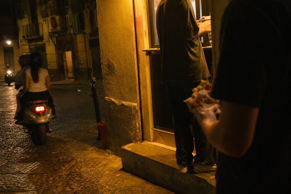 Evening street scene with a lit pizza window casting yellow light on cobbles, locals eating folded pizza in the doorway, scooters passing
