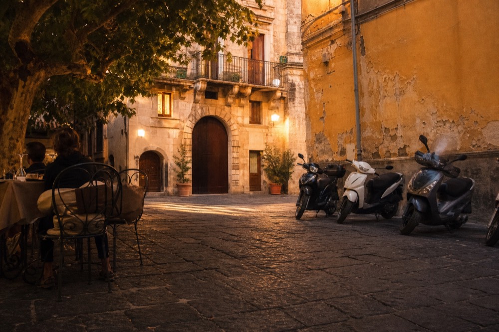 Sunlit square with worn basalt paving, cafe tables under a plane tree, scooters parked along a peeling ochre wall, no signage