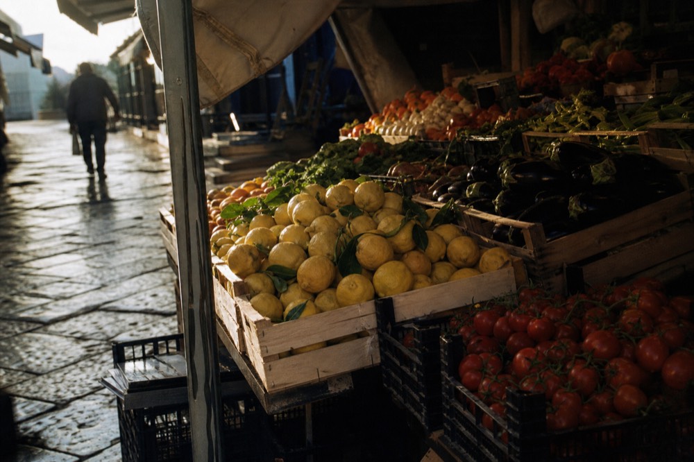 Market stall piled with lemons, tomatoes on the vine and dark aubergines under a canvas awning, crates stacked on wet pavement