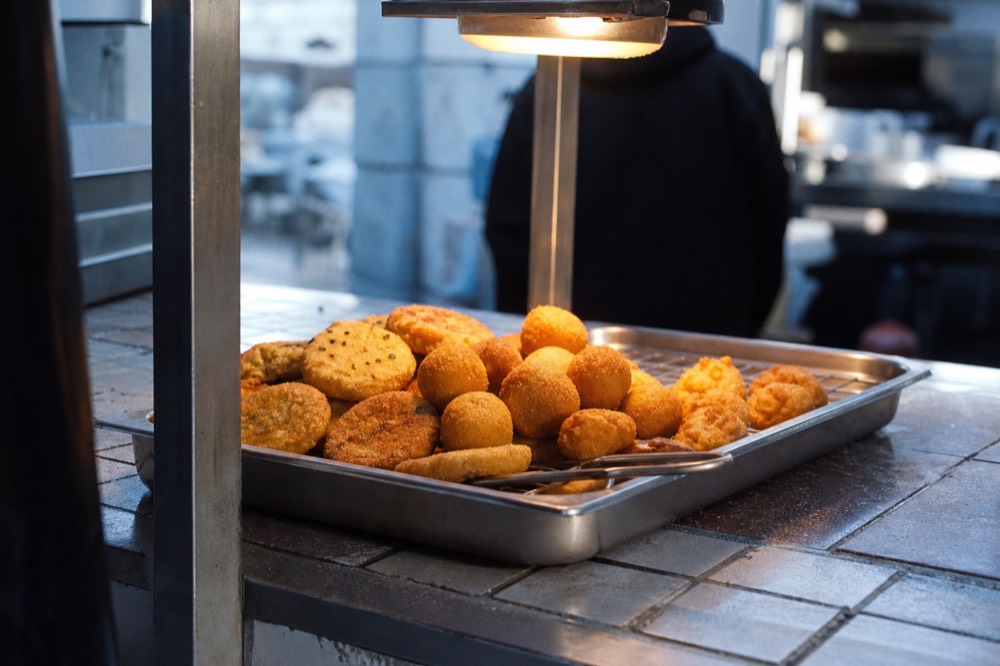 Small round fried pasta cakes and rice croquettes in a metal tray under a heat lamp, tongs resting on the rim, tiled counter