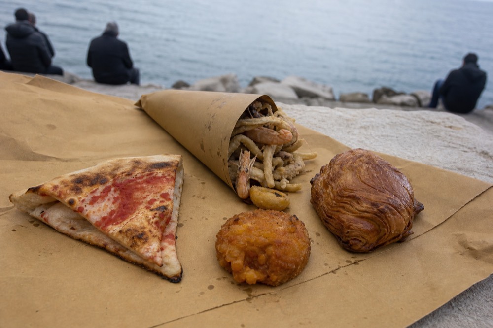 Overhead spread of Neapolitan classics on brown paper: folded pizza slice, paper cone of fried seafood, flaky shell pastry and small fried pasta cake