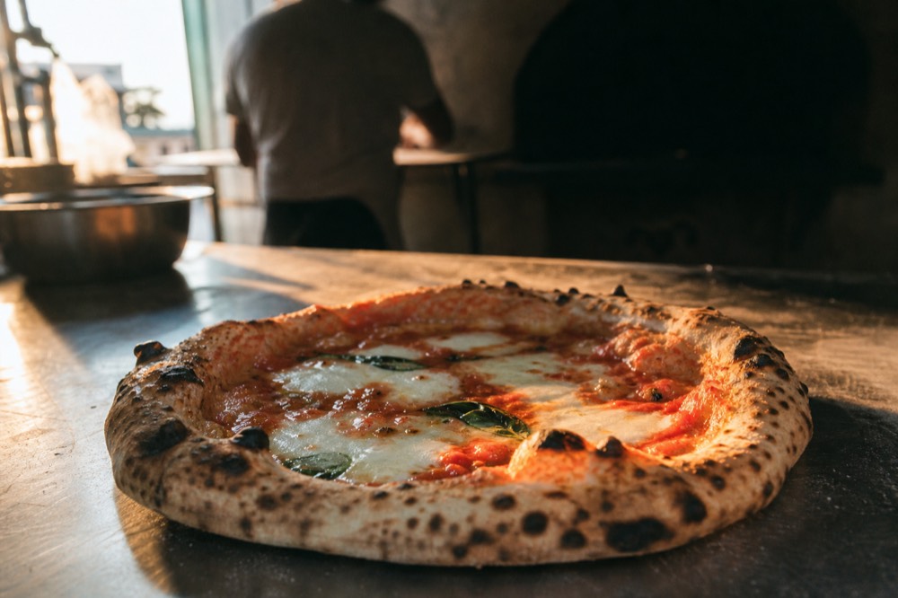 Close-up of a blistered margherita pizza on a metal counter, charred leopard spots on the puffed rim, molten cheese and basil at the soupy center